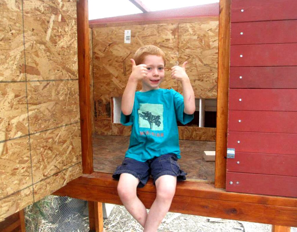 Smiling boy in blue shirt and shorts sits in the henhouse of a chicken coop he helped build